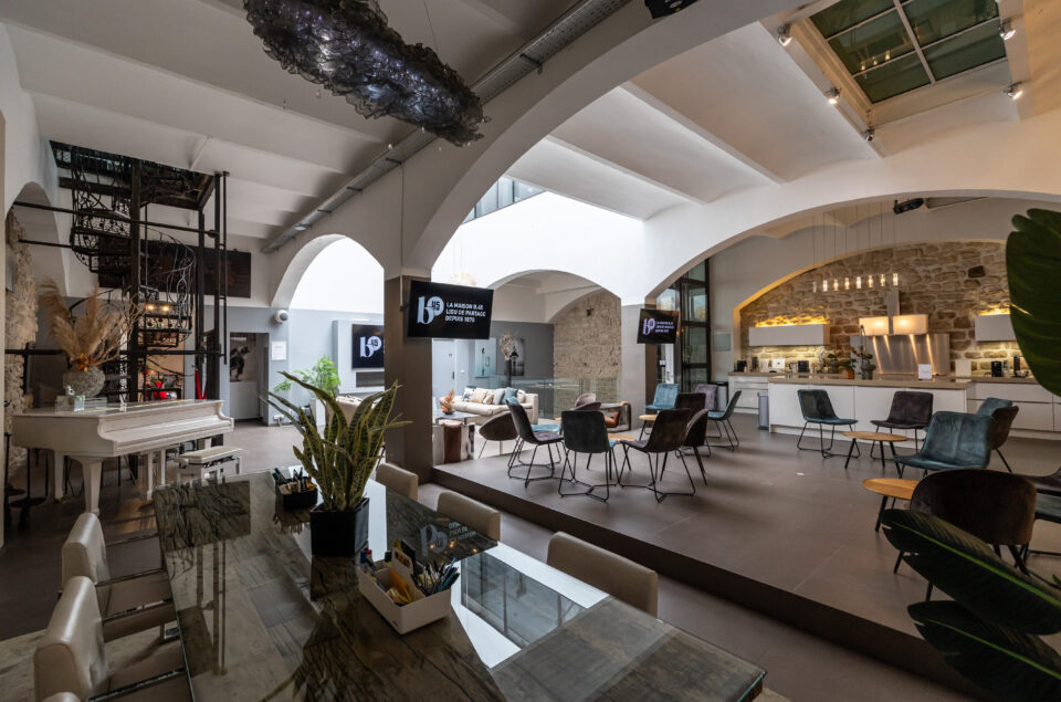 Vision d'angle du grand loft avec escalier en colimaçon sur la gauche et chaises au milieu de la pièce et cuisine sur la droite