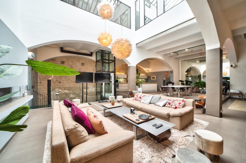 Salle Grand Loft vu d'angle avec grande lumière au plafond effet pompom. Vu des canapés beige avec table basse grise en alu. Arrière-grand-parent plan cuisine du Grand Loft et escalier pour accéder au Lounge