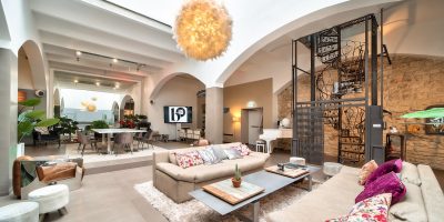 Salle Grand Loft vu d'angle avec grande lumière au plafond effet pompom. Vu des canapés beige avec table basse grise en alu. Arrière-grand-parent plan salle et escalier en colimaçon en fer forgé
