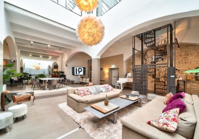 Salle Grand Loft vu d'angle avec grande lumière au plafond effet pompom. Vu des canapés beige avec table basse grise en alu. Arrière-grand-parent plan salle et escalier en colimaçon en fer forgé