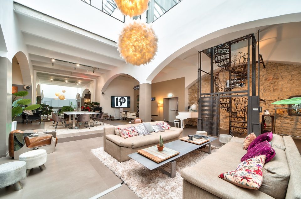 Salle Grand Loft vu d'angle avec grande lumière au plafond effet pompom. Vu des canapés beige avec table basse grise en alu. Arrière-grand-parent plan salle et escalier en colimaçon en fer forgé