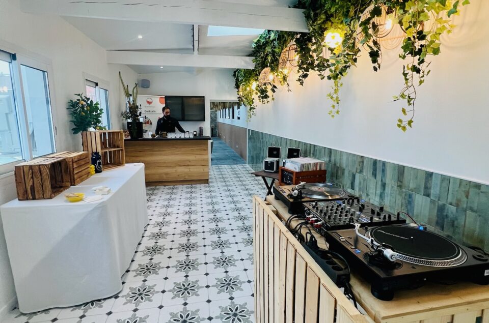Le Salon Alcia. Vision directe du bar en bois avec ses grands fauteuils kaki en velours. Platine à vinyle sur un bar en palette contre le mur. Juste derrière, des vinyles à disposition